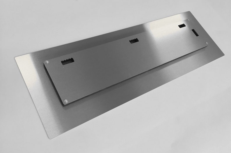 Op de afbeelding: De achterkant van een metalen wandkunstwerk met drie ophanggleuven. Het stuk is zilverkleurig en heeft een geborstelde afwerking.