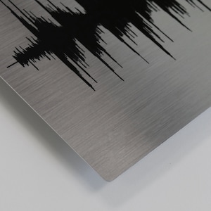 Op de afbeelding: Een close-up van een geborsteld metalen oppervlak met een zwart geluidgolfontwerp. Het metaal heeft een subtiele glans en de geluidgolf is abstract en gestileerd.