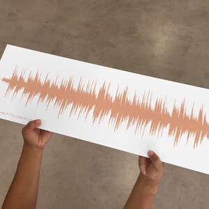 Puede incluir: Una impresi&oacute;n rectangular blanca con un dise&ntilde;o de onda de sonido en un tono naranja apagado. El texto "CAN'T HELP FALLING IN LOVE" est&aacute; impreso en la esquina inferior izquierda. La impresi&oacute;n est&aacute; sostenida por dos manos.