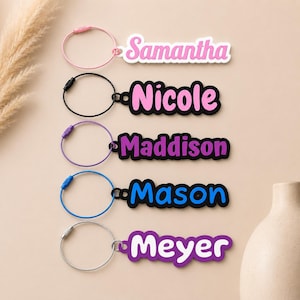 May include: Five custom name keychains with circular wire loops. The names Samantha, Nicole, Maddison, Mason, and Meyer are displayed in colorful, layered script fonts. Each keychain features a distinct color scheme including pink, black, purple, and blue.