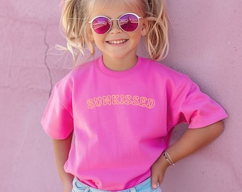 May include: A young girl wearing a pink t-shirt with the text "SUNKISSED" in yellow lettering. She is wearing pink sunglasses and has her hair pulled back in pigtails.
