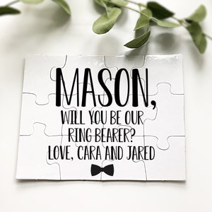 May include: White puzzle with black text that reads "MASON, WILL YOU BE OUR RING BEARER? LOVE, CARA AND JARED" with a black bow tie graphic. The puzzle is made of interlocking pieces.