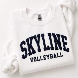 May include: A white crewneck sweatshirt featuring the dark navy blue text SKYLINE in a large arched collegiate font, with the word VOLLEYBALL printed in a smaller, straight navy blue font directly underneath the arched text on the chest.
