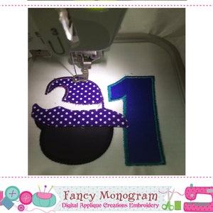 Puede incluir: Una m&aacute;quina de bordar cosiendo un sombrero de bruja morado con lunares blancos y un caldero negro junto a un n&uacute;mero uno azul. El dise&ntilde;o es parte de una creaci&oacute;n de apliques digitales.