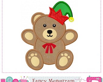 May include: Embroidered teddy bear wearing a red crown and a green elf hat. The bear is brown with tan accents on its paws, ears, and belly. A red bow tie is at the neck. The image includes the text "Fancy Monogram".