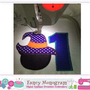 Puede incluir: Una m&aacute;quina de bordar cosiendo un dise&ntilde;o de Halloween. El dise&ntilde;o incluye un sombrero de bruja con lunares morados, una banda naranja y un caldero negro, junto con el n&uacute;mero 1 en azul. El texto "Fancy Monogram" es visible.
