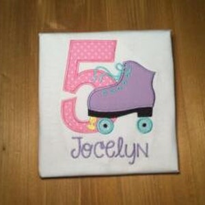 May include: A white t-shirt with a pink number 5 and a purple roller skate with blue wheels. The name Jocelyn is embroidered below the skate.