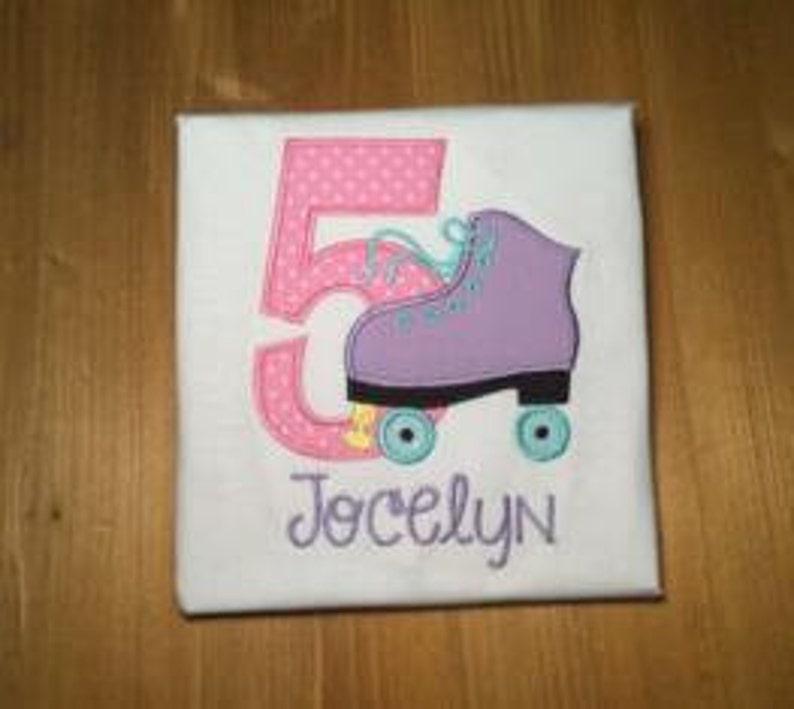 May include: A white t-shirt with a pink number 5 and a purple roller skate with blue wheels. The name Jocelyn is embroidered below the skate.