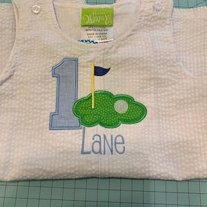 May include: White seersucker romper with a blue gingham number 1 and the name "Lane" embroidered in blue. Features a green golf course design with a golf ball and flag. Includes snap closures.