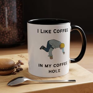 May include: White ceramic coffee mug with a black interior and handle. The mug features the text "I LIKE COFFEE IN MY COFFEE HOLE" with a cartoon illustration of a person bending over. A spoon and a macaron are in the foreground.