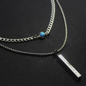 May include: A silver chain necklace with a blue gemstone pendant and a silver bar pendant. The bar pendant has a small inscription.