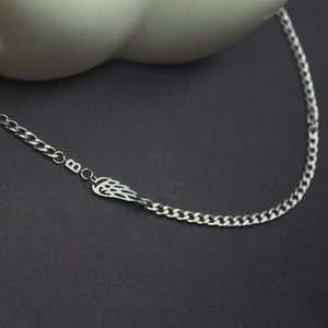 Può includere: Una collana a catena color argento con un ciondolo a forma di ala. La collana ha un design a catena a maglia e viene presentata su uno sfondo scuro. Il ciondolo ad ala è un design ritagliato.
