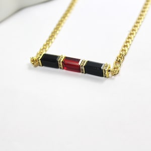 Puede incluir: Un collar de cadena dorada con un colgante que presenta tres piedras rectangulares: dos negras y una roja.