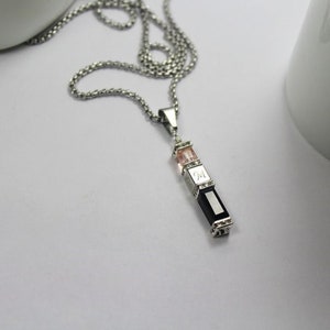 May include: A silver chain necklace with a pendant featuring three rectangular stones. The top stone is pink, the middle stone is engraved with the letter "M", and the bottom stone is black.