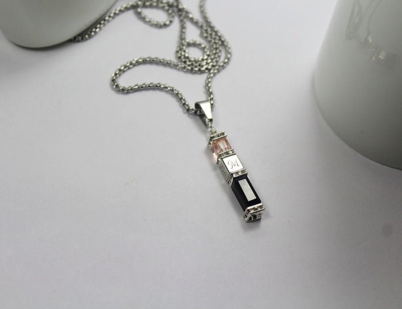 May include: A silver chain necklace with a pendant featuring three rectangular stones. The top stone is pink, the middle stone is engraved with the letter "M", and the bottom stone is black.