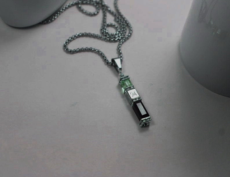 May include: A silver chain necklace with a pendant featuring three rectangular stones. The top stone is green, the middle stone is silver with the letter "M" engraved on it, and the bottom stone is dark purple.