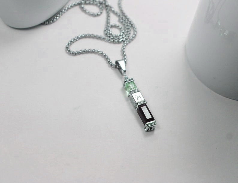 May include: A silver chain necklace with a pendant featuring three rectangular stones. The top two stones are green, and the bottom stone is dark purple. The pendant has a small silver charm with the letter "M" engraved on it.
