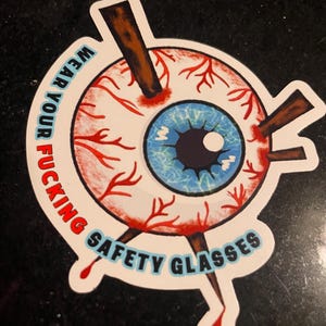 Può includere: Un adesivo con la grafica di un occhio trafitto da paletti di legno. L'occhio ha un'iride blu e vene rosse. Il testo intorno all'occhio recita "WEAR YOUR FUCKING SAFETY GLASSES" in grassetto.