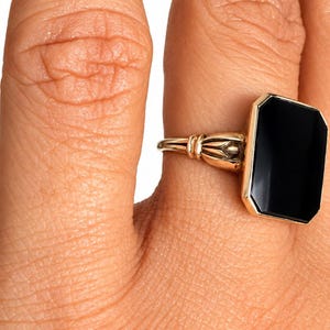 May include: A gold ring featuring a rectangular black stone set in a polished metal frame. The band displays a decorative, ribbed design where it connects to the setting, providing a structured look to the piece of jewelry.