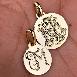 May include: Two circular gold pendants with polished surfaces. The larger pendant features an intricate, overlapping monogram of the letters M and N. The smaller pendant displays a single, elegant script letter M engraved in the center.