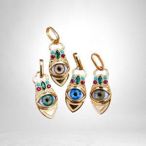 May include: Four gold-toned pendants shaped like eyes, each featuring a central iris in blue, brown, or green. The pendants are accented with small turquoise, emerald green, and ruby red stones arranged above the eye, with a metal bail at the top.