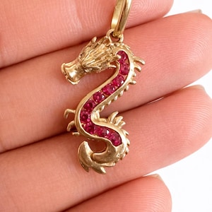 May include: A gold dragon pendant featuring a curved body set with a series of small, round, vibrant red gemstones along its spine. The pendant includes a polished gold bail at the top for attachment to a chain, showcasing detailed scales and features.