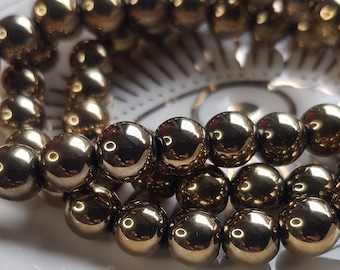 May include: A close-up of a bracelet made of shiny, metallic gold beads. The beads are round and smooth, and they are arranged in a single row.