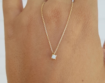 May include: A delicate gold chain necklace featuring a single, small, round, clear faceted gemstone pendant held in a four-prong setting. The necklace is composed of a thin, polished gold link chain that rests against the skin, highlighting the sparkle.
