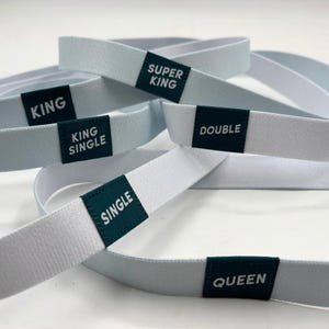 May include: Several light blue elastic bands with dark green labels. The labels read "KING", "SUPER KING", "KING SINGLE", "DOUBLE", "SINGLE", and "QUEEN". The bands are arranged in a layered pattern.