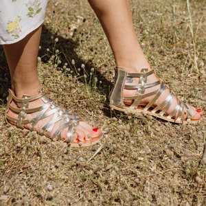 Op de afbeelding: Een paar gouden gladiatorsandalen met banden en gespen. De sandalen worden gedragen door iemand die op een grasveld staat.