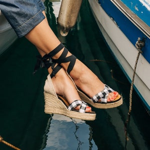 Könnte beinhalten: Eine Person in Jeans und schwarz-weiß karierten Keilsandalen mit schwarzen Schleifen sitzt am Rand eines weißen Bootes. Das Boot liegt neben einem blauen Boot mit einem weißen Streifen. Das Wasser ist grün und im Vordergrund befindet sich ein Seil.