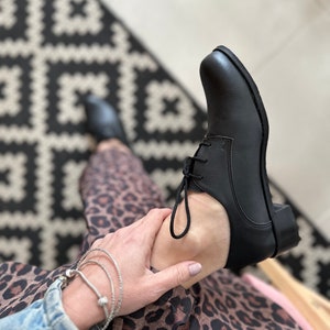Op de afbeelding: Zwarte leren veterschoenen met een lage hak. De schoenen worden gedragen door iemand die een luipaardprintrok en een spijkerjack draagt.