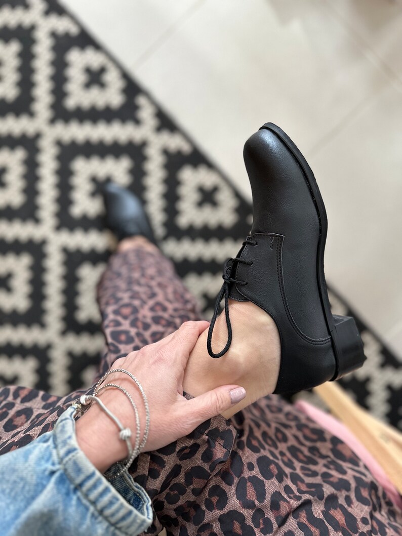 Op de afbeelding: Zwarte leren veterschoenen met een lage hak. De schoenen worden gedragen door iemand die een luipaardprintrok en een spijkerjack draagt.
