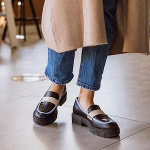 May include: A pair of black leather loafers with a light blue trim and a white stripe across the top. The loafers have a chunky platform sole and are worn with blue jeans.