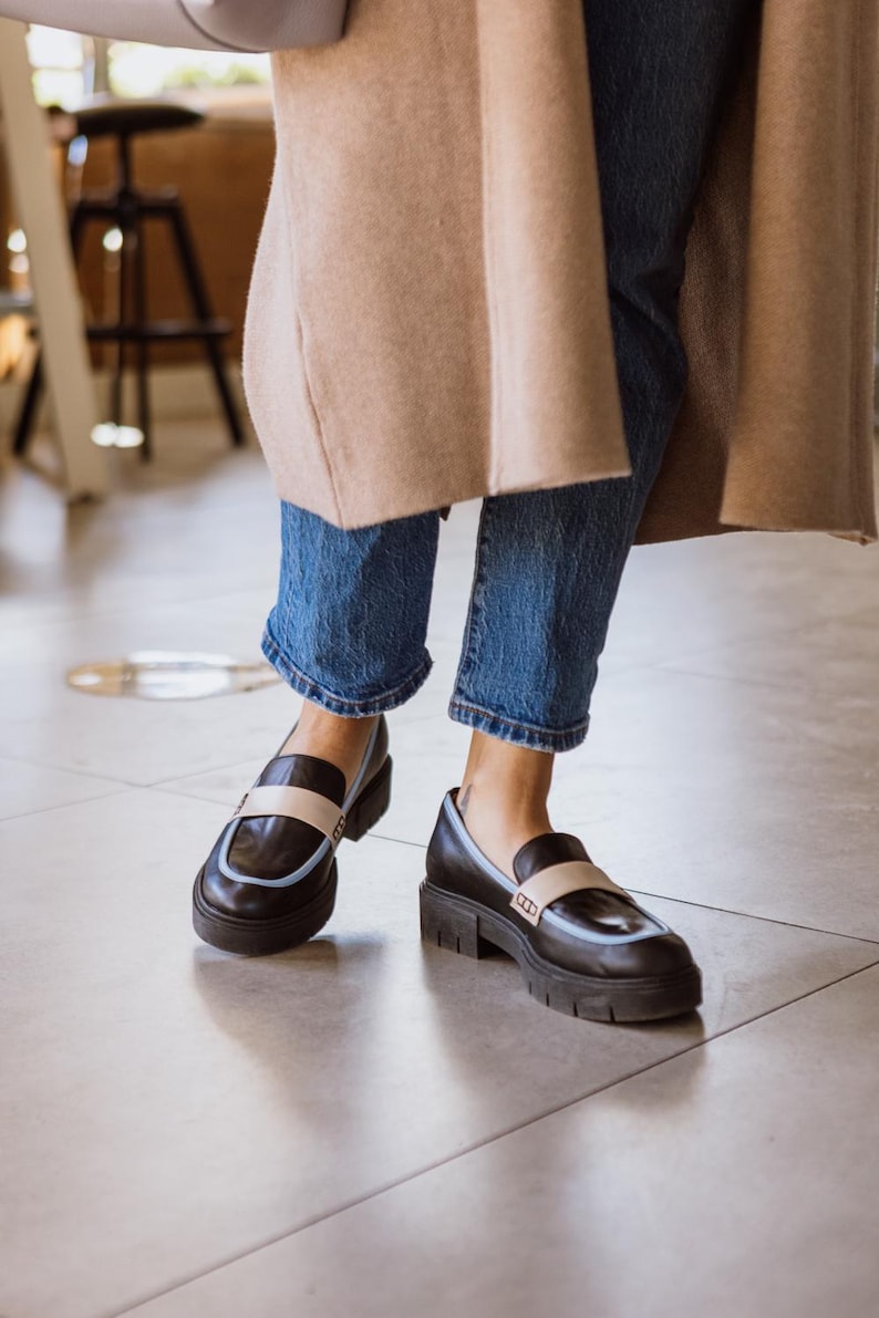 May include: A pair of black leather loafers with a light blue trim and a white stripe across the top. The loafers have a chunky platform sole and are worn with blue jeans.
