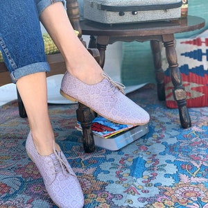 Pu&ograve; includere: Un paio di scarpe con lacci in pelle color lilla chiaro con un motivo testurizzato. Le scarpe sono indossate da una persona seduta su una sedia con le gambe incrociate.