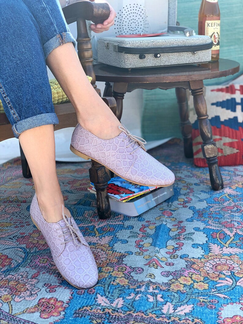 Pu&ograve; includere: Un paio di scarpe con lacci in pelle color lilla chiaro con un motivo testurizzato. Le scarpe sono indossate da una persona seduta su una sedia con le gambe incrociate.