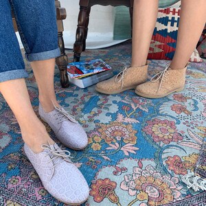 Pu&ograve; includere: Due paia di scarpe con lacci per donna. Un paio &egrave; rosa chiaro con un motivo testurizzato e l'altro paio &egrave; beige con una texture simile al sughero. Le scarpe sono su un tappeto floreale colorato.
