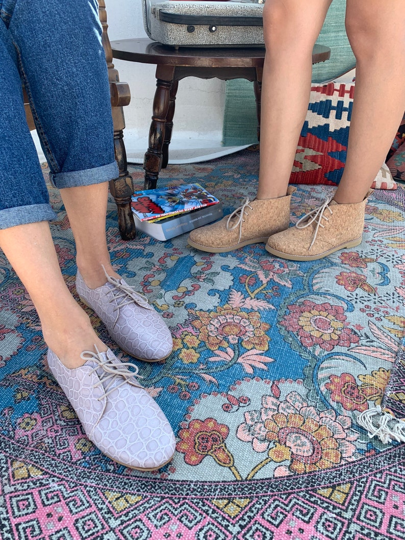 Pu&ograve; includere: Due paia di scarpe con lacci per donna. Un paio &egrave; rosa chiaro con un motivo testurizzato e l'altro paio &egrave; beige con una texture simile al sughero. Le scarpe sono su un tappeto floreale colorato.