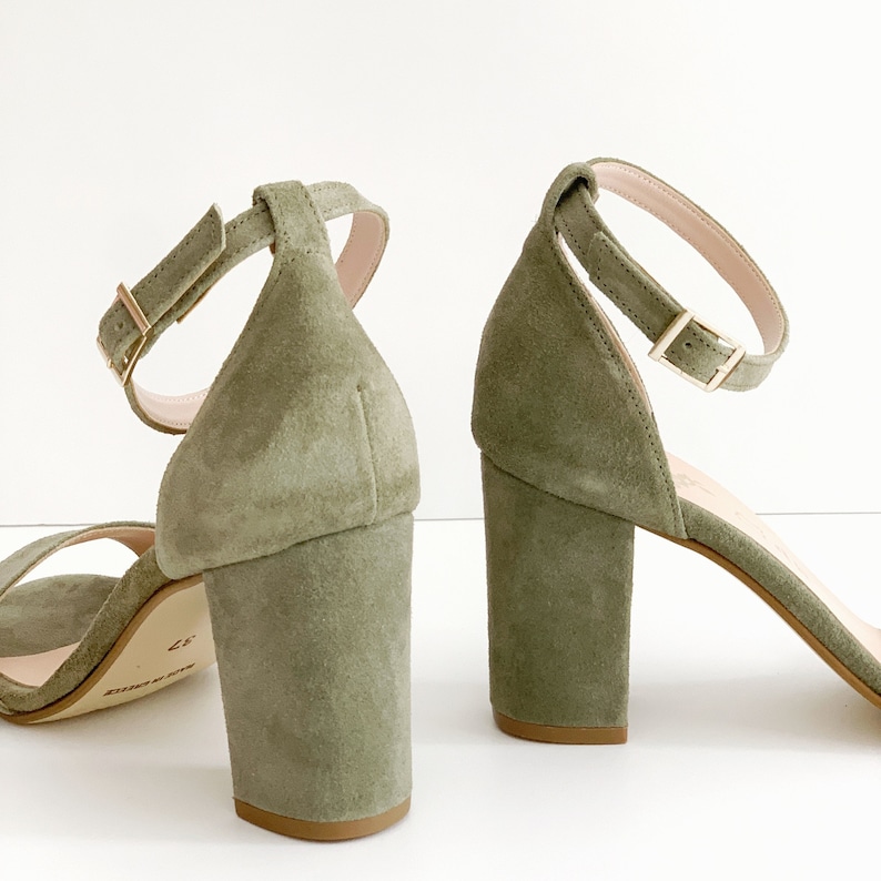 May include: A pair of green suede heeled sandals with ankle straps and gold buckles. The heels are block heels.