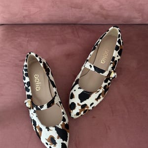 May include: A pair of white and brown leopard print shoes with a black strap and gold buckle. The shoes are pointed toe flats with the brand name "aelia" written on the inside of each shoe.