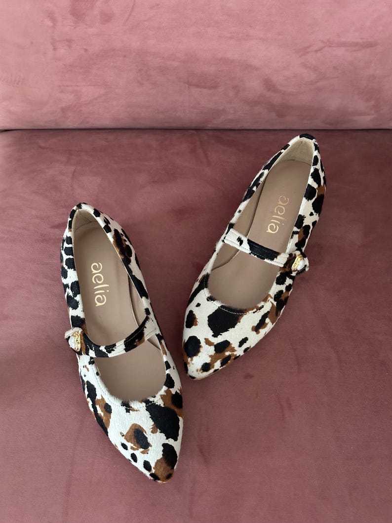May include: A pair of white and brown leopard print shoes with a black strap and gold buckle. The shoes are pointed toe flats with the brand name "aelia" written on the inside of each shoe.