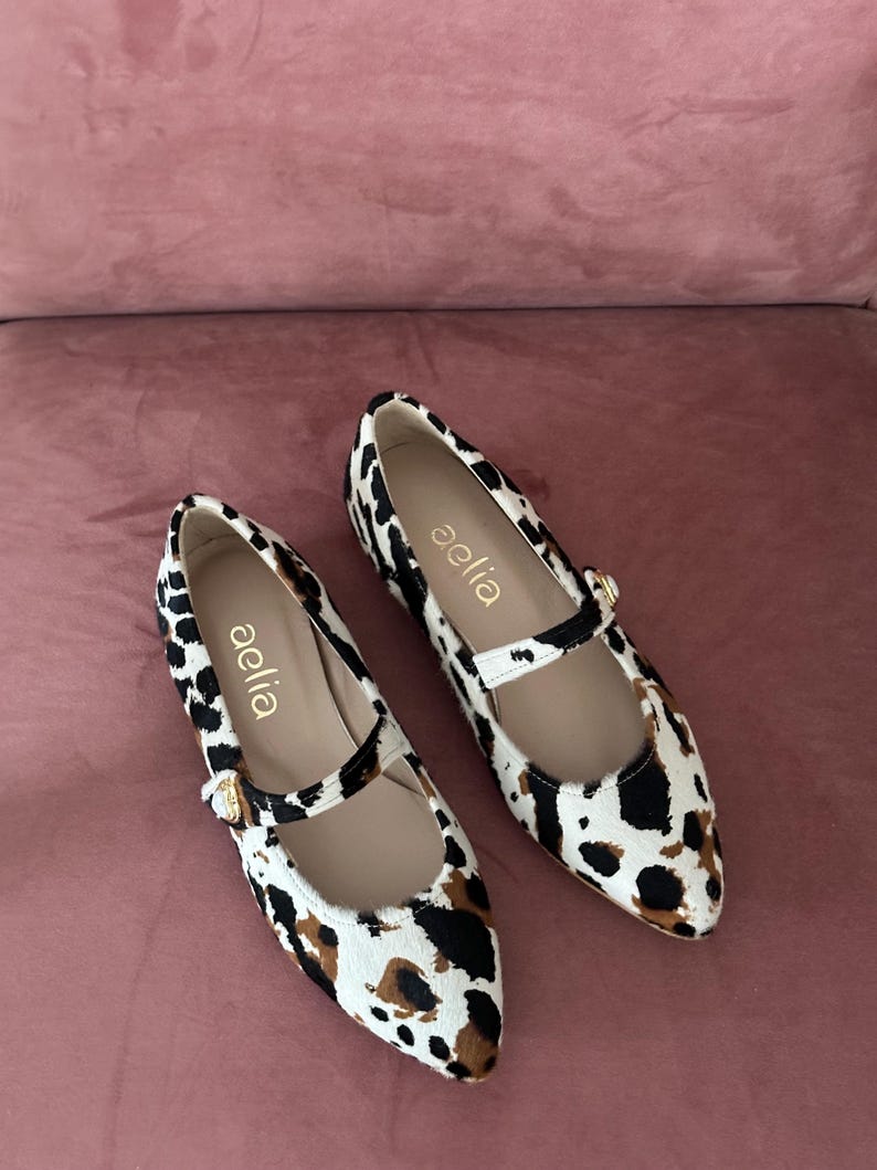 May include: A pair of white and brown leopard print ballet flats with a gold buckle and the word "aelia" written on the side.