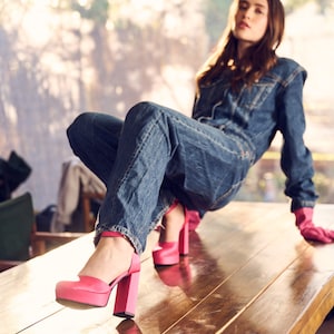 Puede incluir: Una persona con un mono vaquero y tacones de plataforma rosa se sienta en una mesa de madera. Las piernas de la persona est&aacute;n cruzadas y su mano derecha est&aacute; apoyada en la mesa.