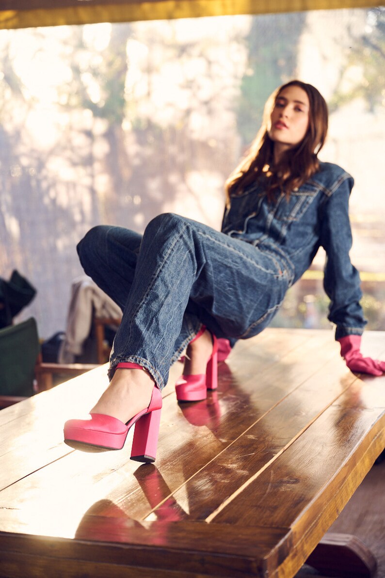 Puede incluir: Una persona con un mono vaquero y tacones de plataforma rosa se sienta en una mesa de madera. Las piernas de la persona est&aacute;n cruzadas y su mano derecha est&aacute; apoyada en la mesa.