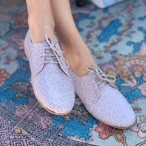 Pu&ograve; includere: Un paio di scarpe con lacci in pelle color lilla chiaro con un motivo testurizzato. Le scarpe sono su un tappeto colorato.