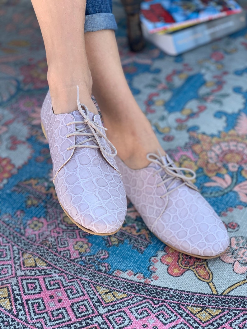Pu&ograve; includere: Un paio di scarpe con lacci in pelle color lilla chiaro con un motivo testurizzato. Le scarpe sono su un tappeto colorato.