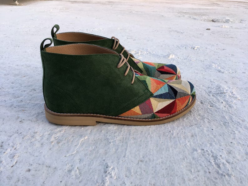 May include: Green suede ankle boots with a colourful geometric pattern on the toe and heel. The boots have a lace-up closure and a brown sole.