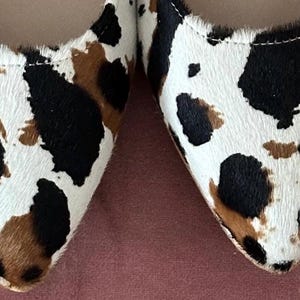 May include: A pair of pointed-toe flats with a cow print pattern in shades of white, brown, and black.