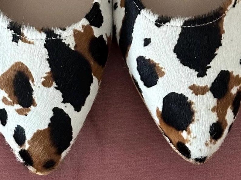 May include: A pair of pointed-toe flats with a cow print pattern in shades of white, brown, and black.
